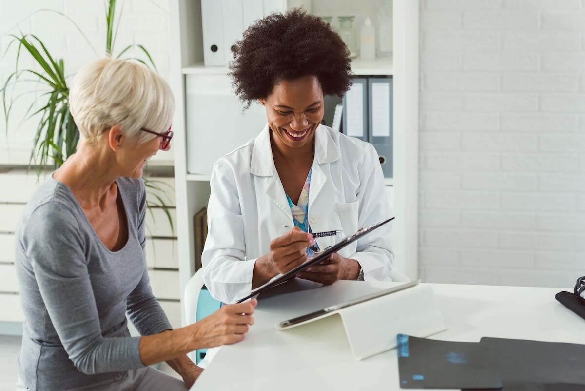 Smiling healthcare provider in white coat writing notes while consulting with older patient in gray top in bright medical office