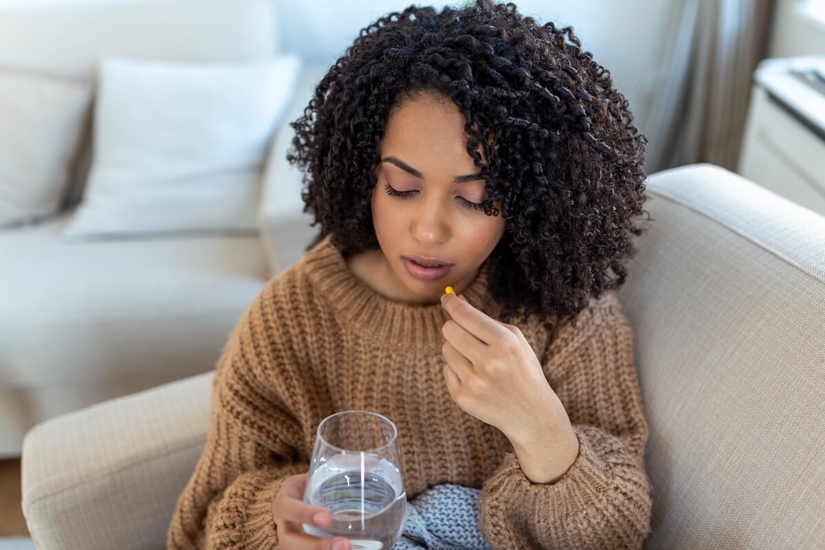 Person with curly hair in brown sweater taking medication with water, sitting on light-colored couch in home setting