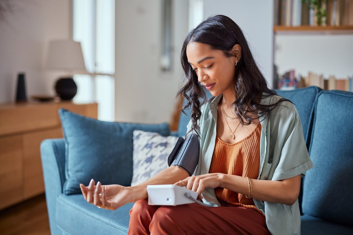 Woman in sage shirt checking blood pressure monitor while sitting on blue couch at home