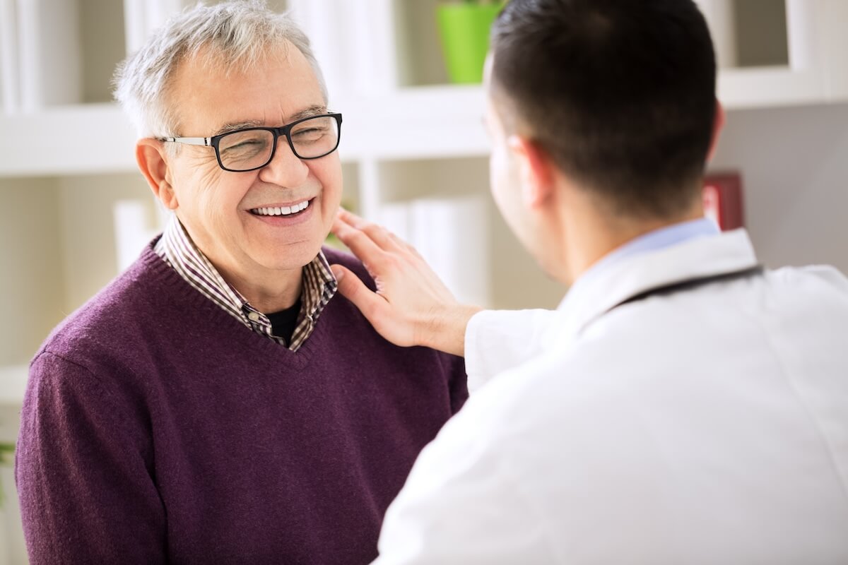 Smiling older patient in purple sweater receiving reassuring touch from healthcare provider in white coat during medical consultation