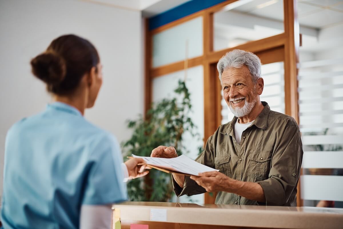Senior patient exchanging paperwork with healthcare worker at medical office reception desk with wooden partitions and plants in background