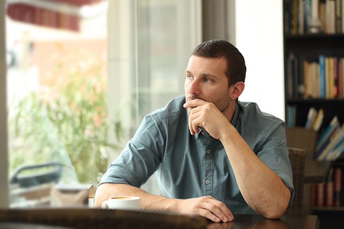 Person in blue shirt looking pensively out window, hand on chin, with coffee cup on table and bookshelf in background
