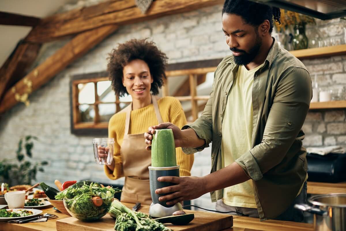 Two people making green smoothies in rustic kitchen with brick walls, wooden counter filled with fresh vegetables and salad ingredients