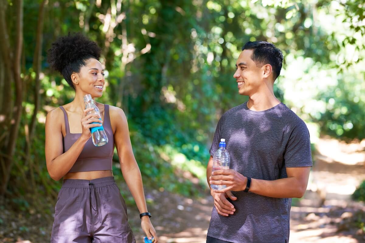 Two people smiling at each other on a wooded trail, holding water bottles and wearing athletic clothing during an outdoor workout.
