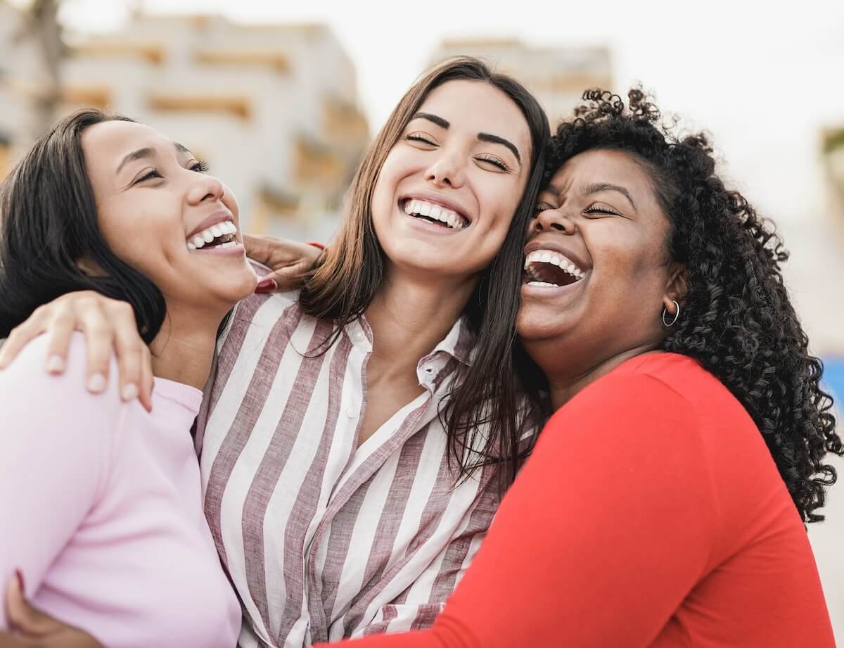 Three women laughing together, arms around each other, wearing pink, striped, and red tops