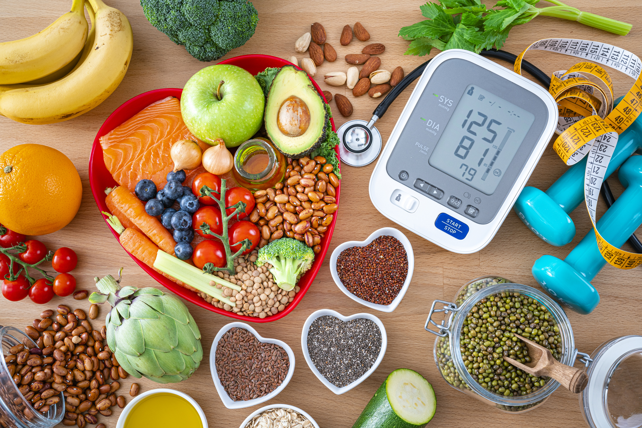 Heart-healthy foods arranged with blood pressure monitor, dumbbells and measuring tape. Heart-shaped dish contains salmon, fruits, vegetables and legumes