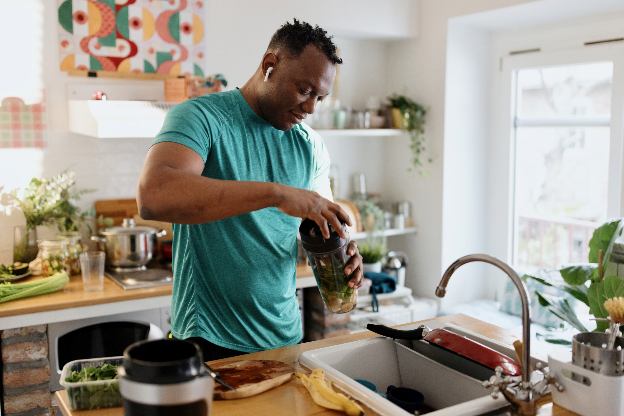 Person in teal shirt making smoothie in bright kitchen. Wearing earbuds while preparing healthy ingredients with plants and colorful art nearby