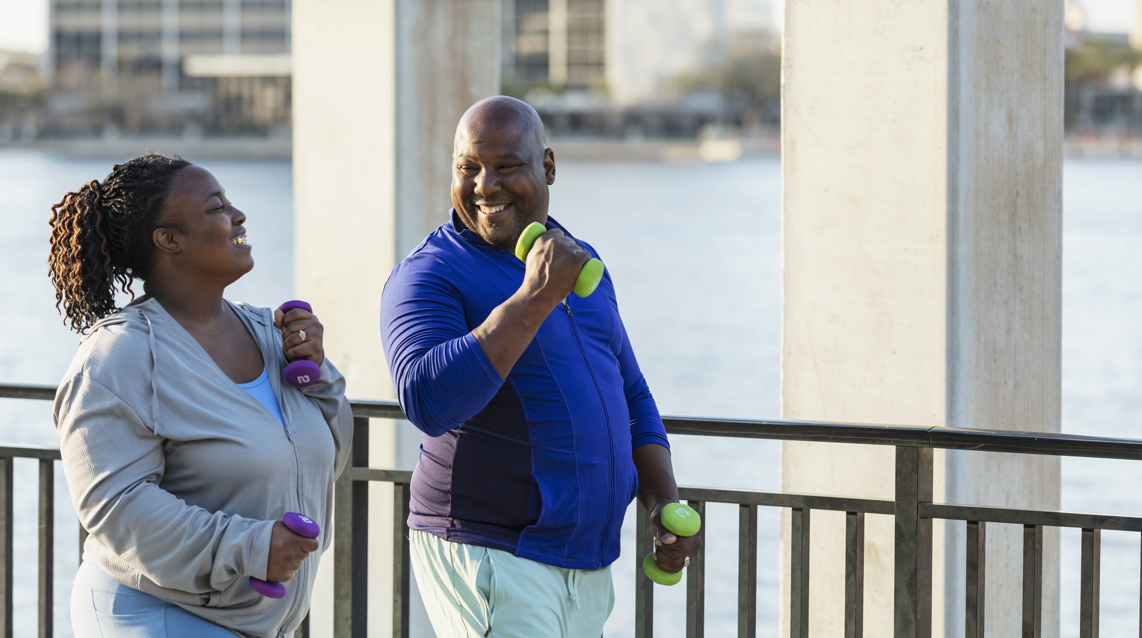 Two people exercising with colorful dumbbells by waterfront. Smiling while working out together on boardwalk with urban skyline background