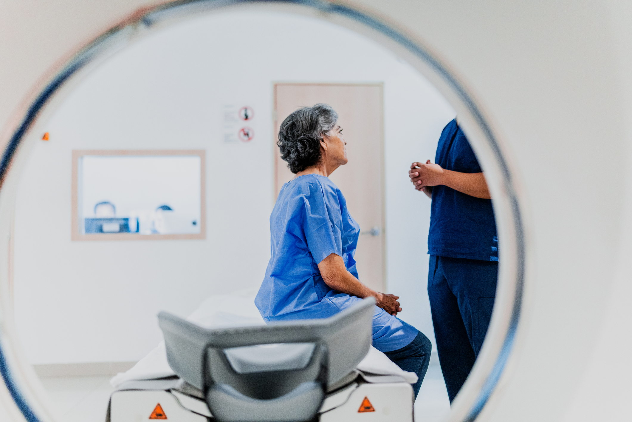 Patient in blue gown sitting on imaging equipment with medical professional nearby. View through CT or MRI scanner in clinical setting