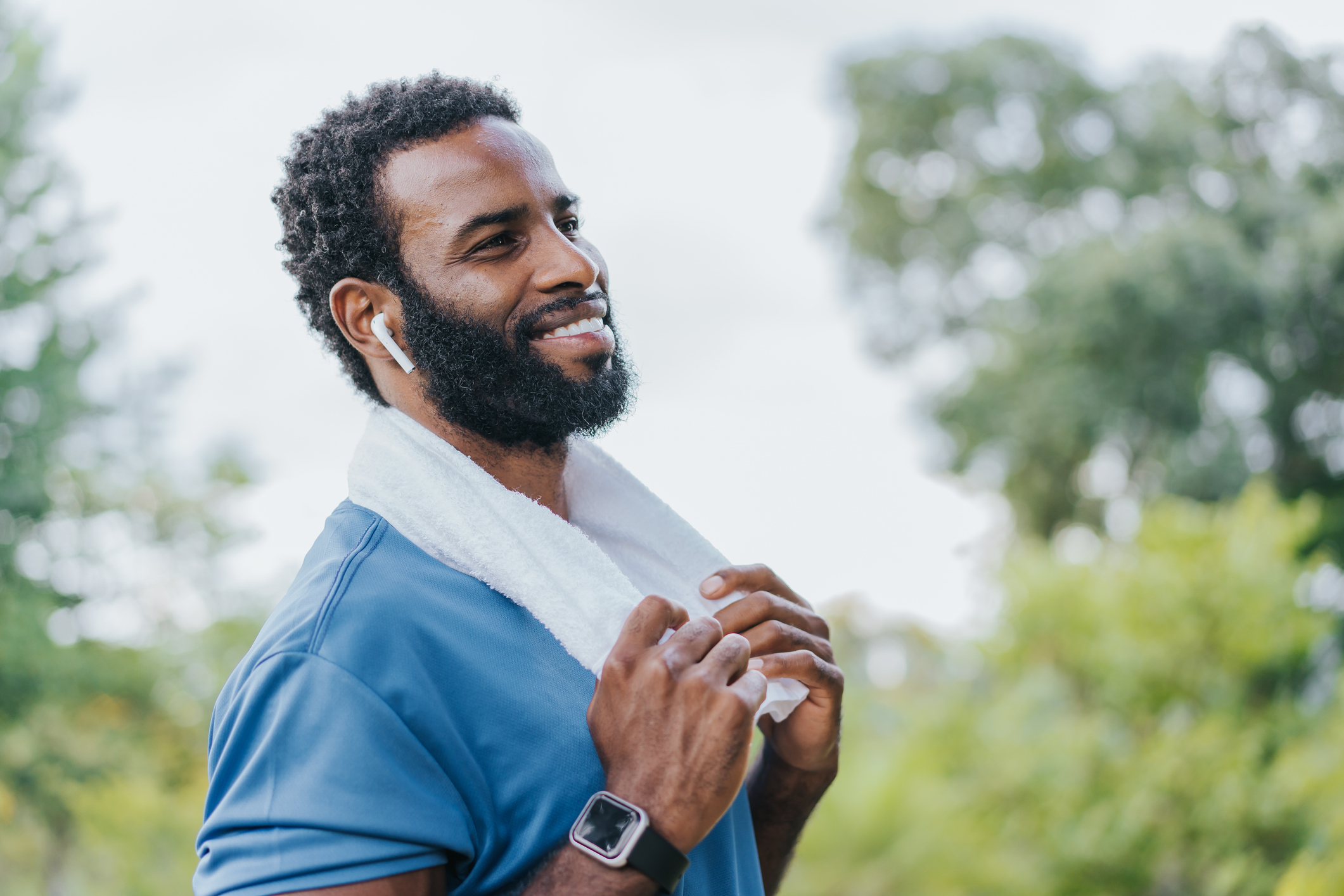 Smiling man with beard wearing blue athletic shirt and white towel around neck. He has wireless earbuds and a smartwatch, enjoying time outdoors.