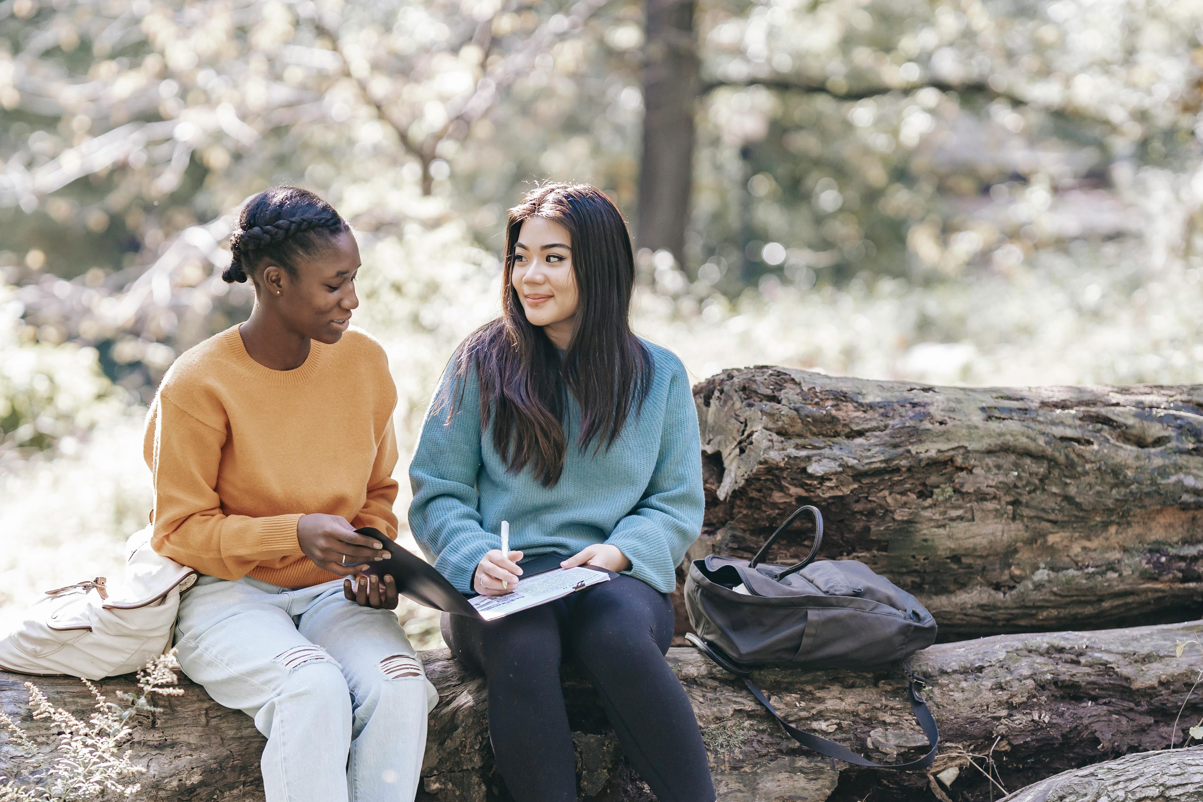 Two students studying on fallen log in forest. One in yellow sweater, one in blue, taking notes with backpacks nearby