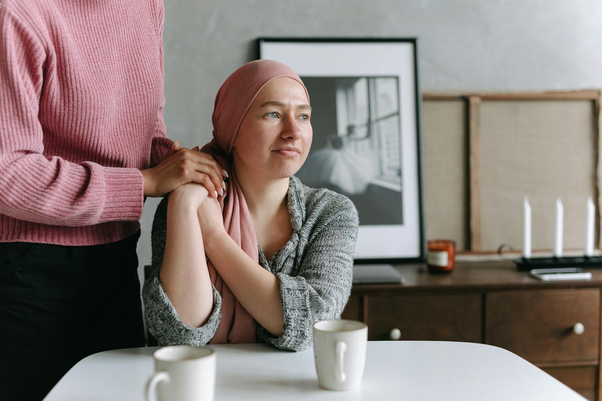 Woman in pink headscarf at table with supportive person's hand on shoulder in indoor setting