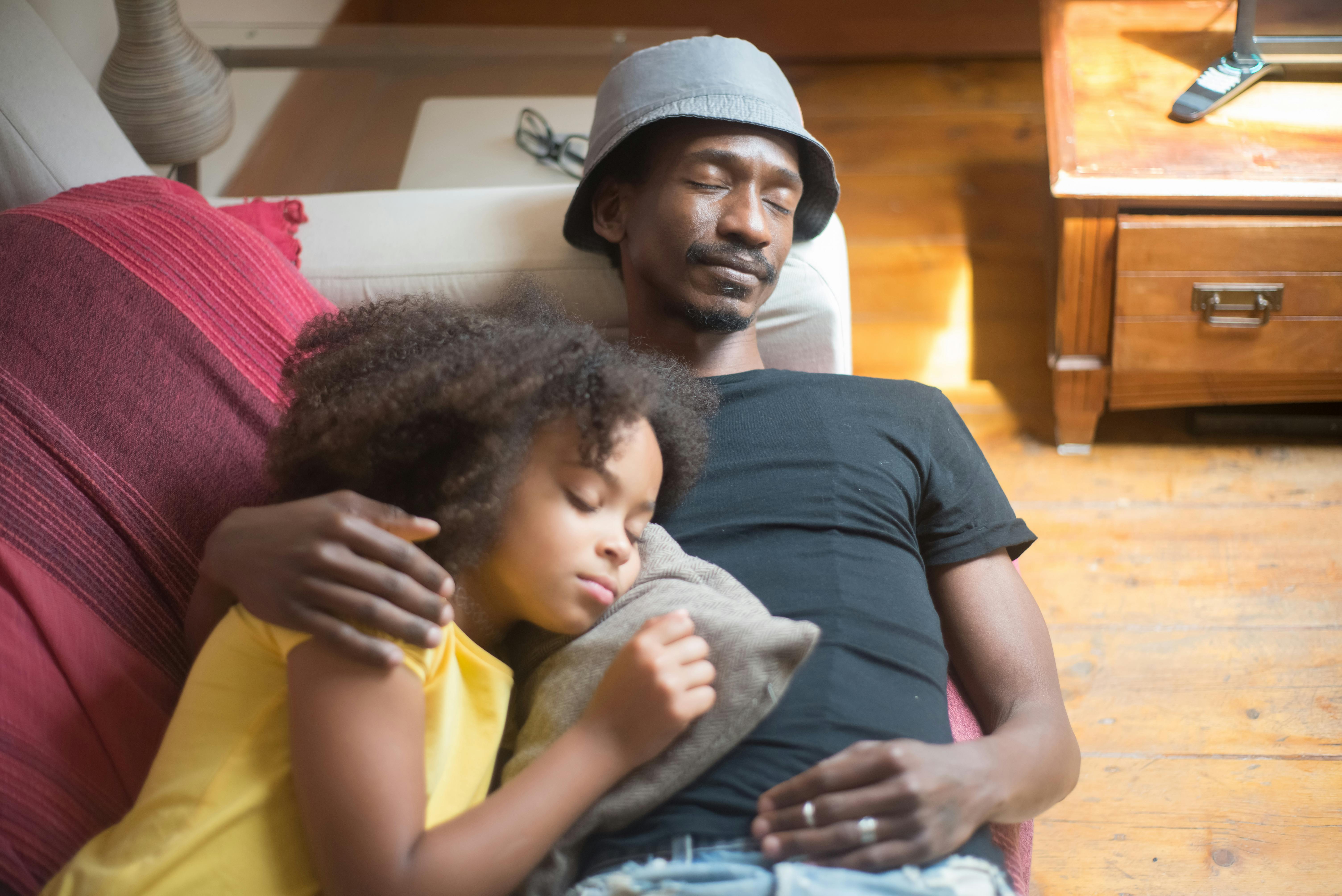 Father wearing light blue cap holding young child in yellow shirt on burgundy couch in home interior with wooden furniture