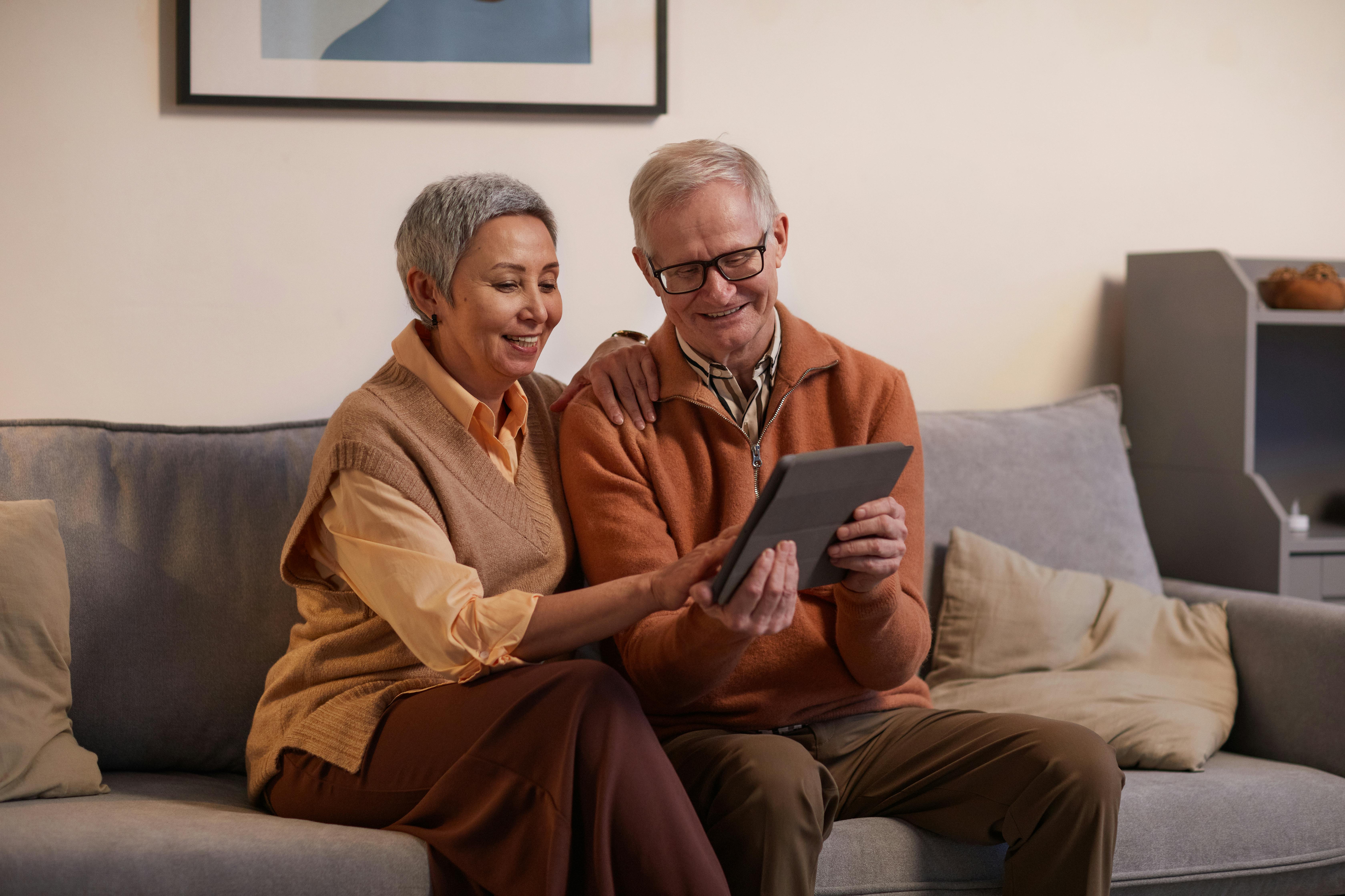 Older couple sitting together on gray couch smiling while using tablet device in modern living room with beige walls