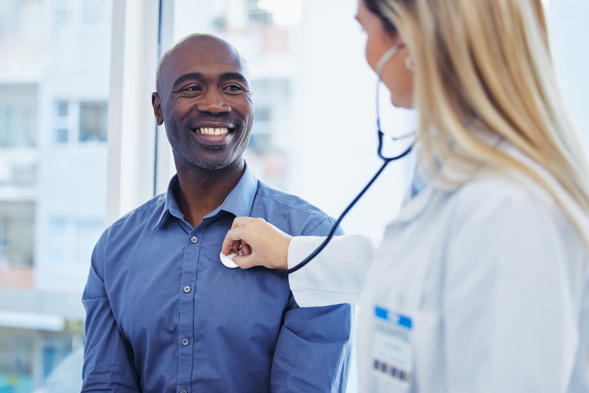 Female doctor examining smiling male patient with stethoscope in modern medical office with city view