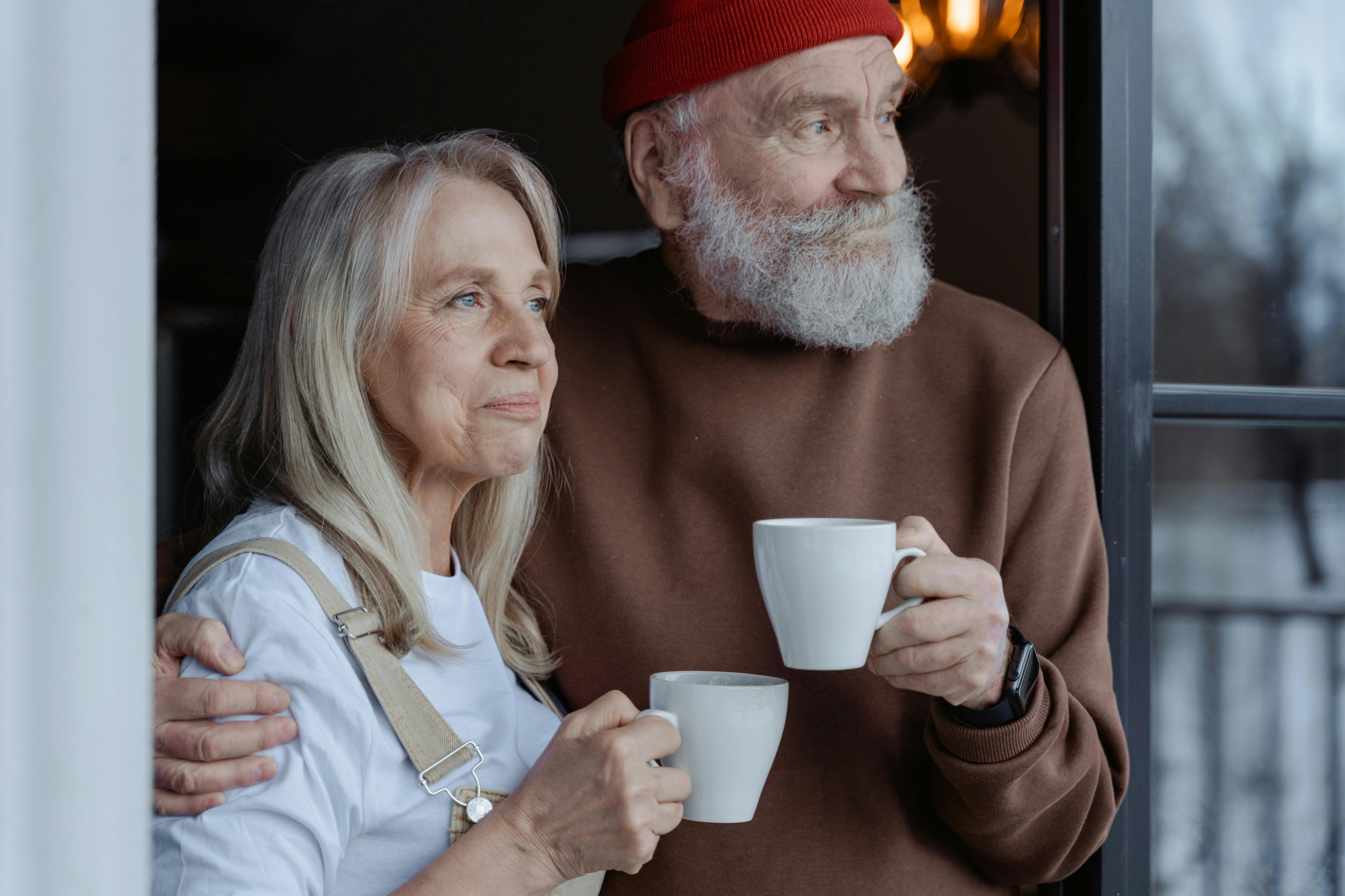 Elderly couple holding white mugs at doorway, woman in blue shirt and man in red beanie and brown sweater