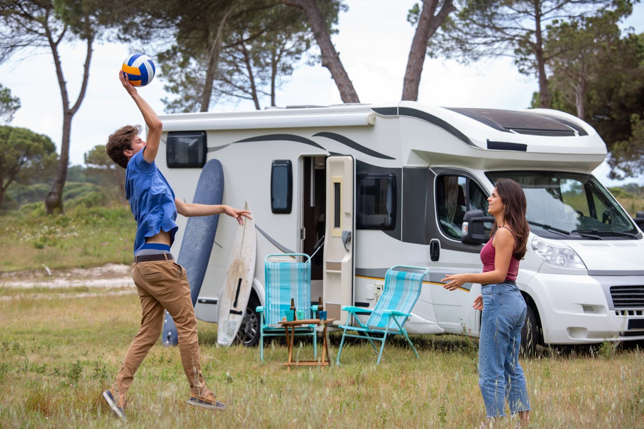 Boy playing volleyball with girl outside white RV camper van in natural outdoor setting with trees
