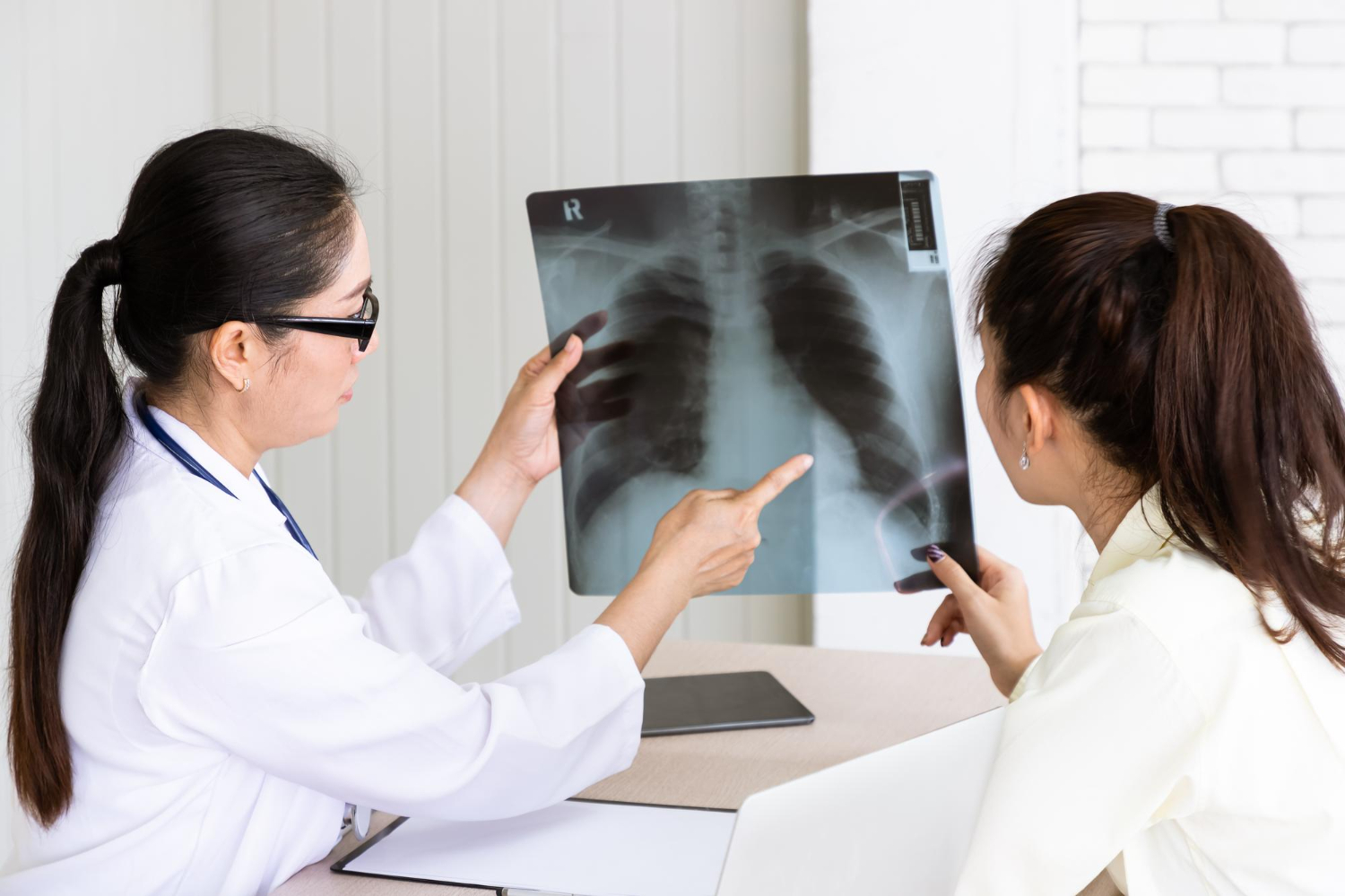 Female doctor in white coat and glasses explaining chest X-ray to patient at desk in medical office