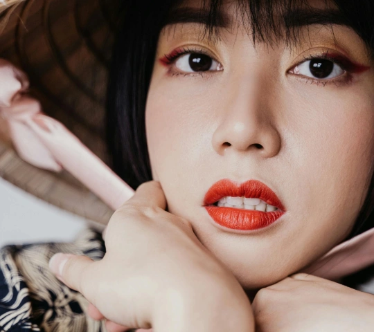 Close-up of a woman with black hair wearing red lipstick and winged eyeliner, holding a pink ribbon near her face.