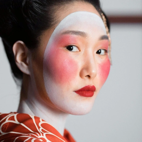 Portrait of a woman with traditional Geisha-style makeup, including white face paint, red cheeks, and red lipstick, wearing an orange patterned garment.