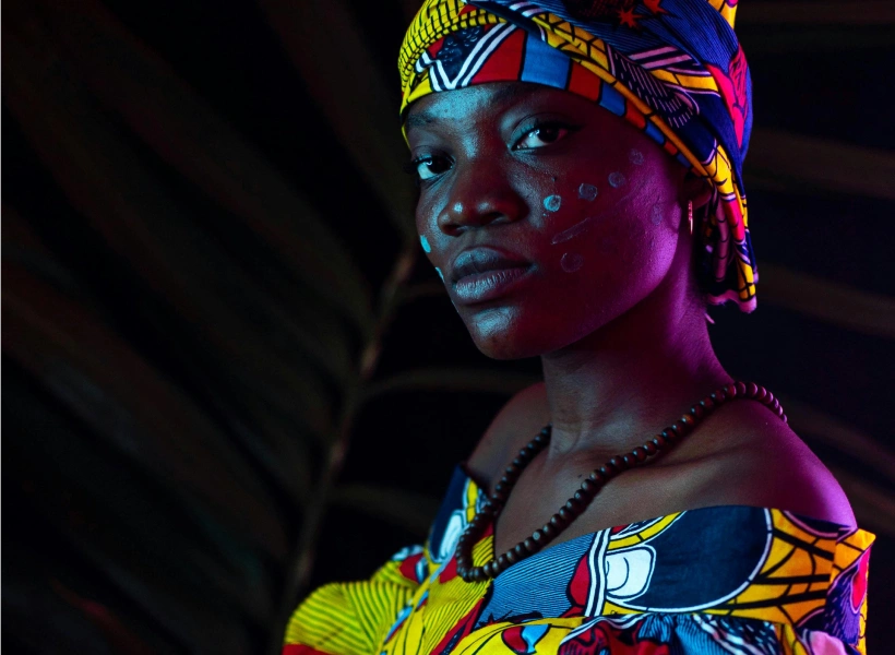 Portrait of a woman wearing a colorful patterned headwrap and dress with white face paint dots on her cheek and a beaded necklace.