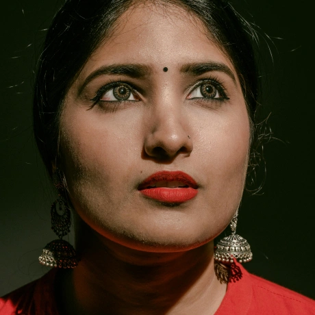 Close-up of a woman with red lipstick, wearing traditional silver jhumka earrings and a black bindi, looking slightly upward against a dark background.