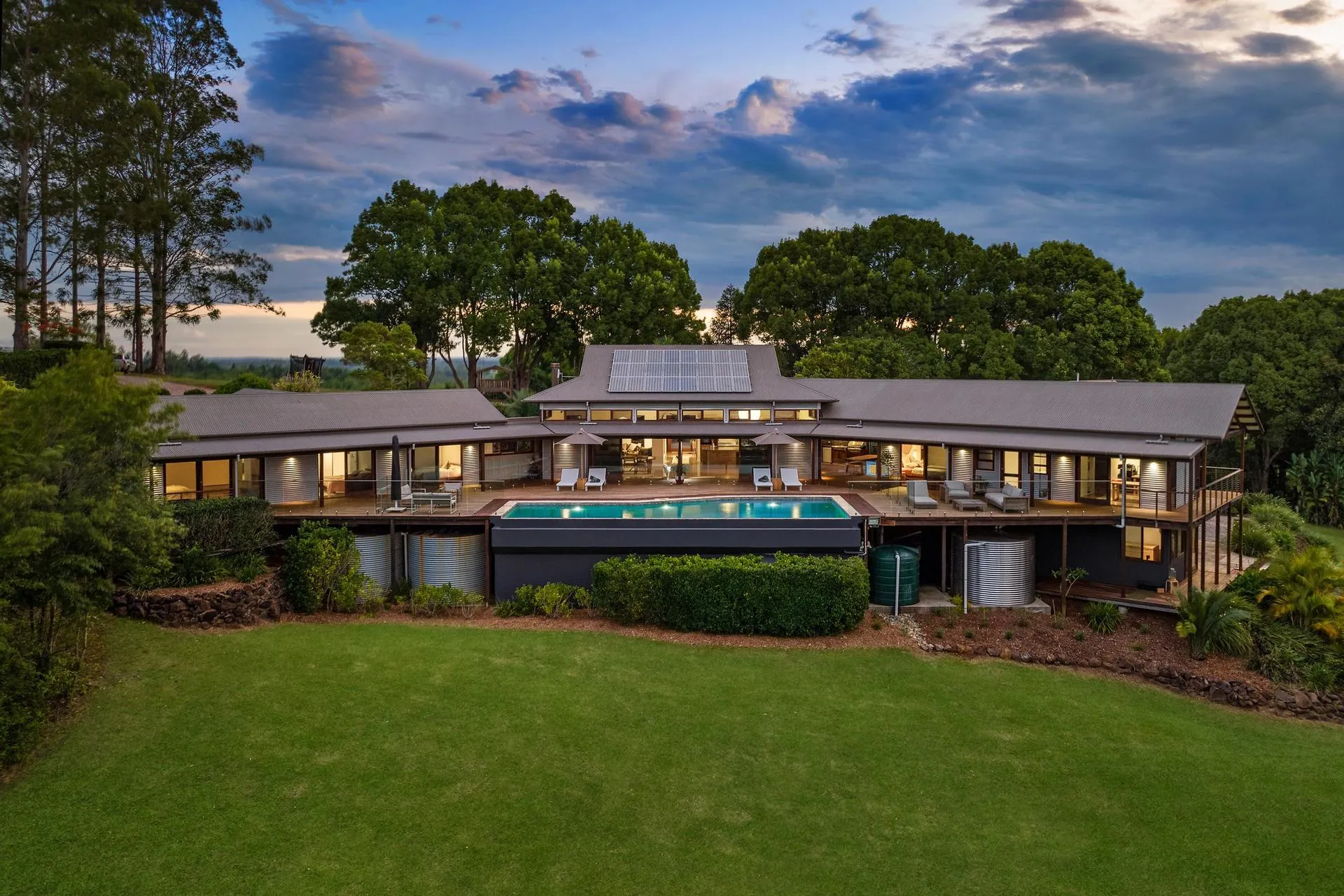 Modern house with solar panels on the roof, large outdoor swimming pool, surrounded by lush green trees and lawn at dusk.