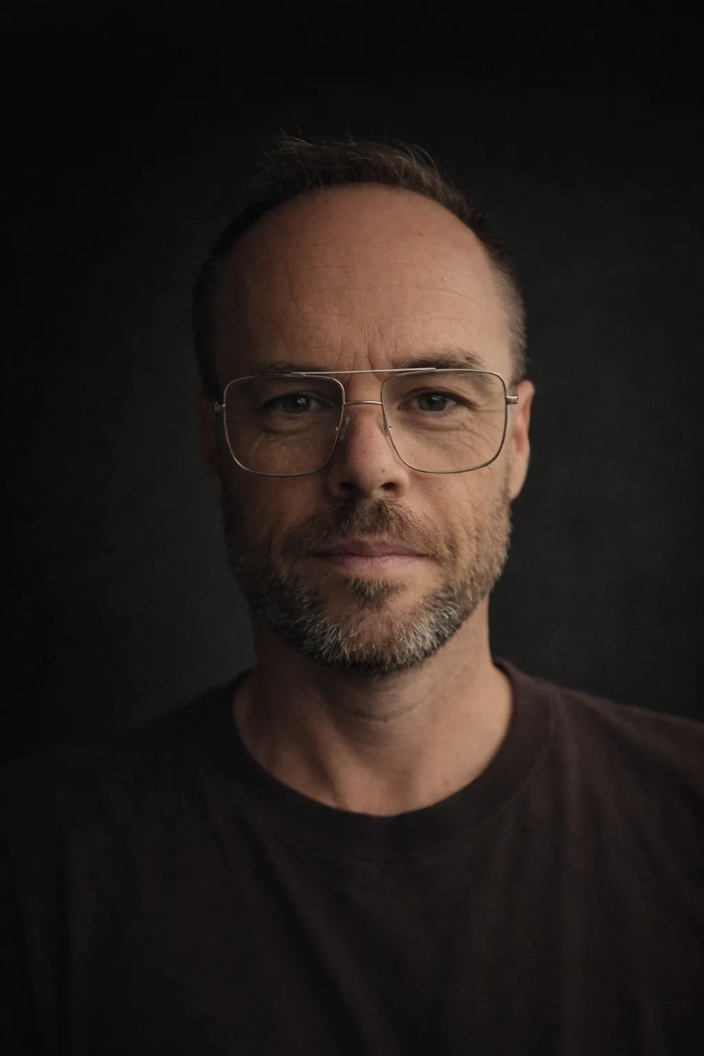 Sam McCaig Man with short hair, beard, and large glasses wearing a dark shirt, against a dark background.