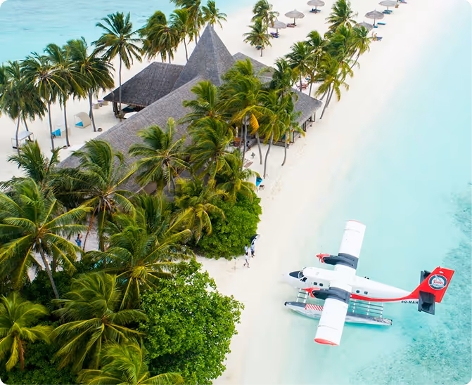 Aerial view of a tropical beach resort with palm trees, thatched-roof buildings, beach umbrellas, and a seaplane docked on clear turquoise water.
