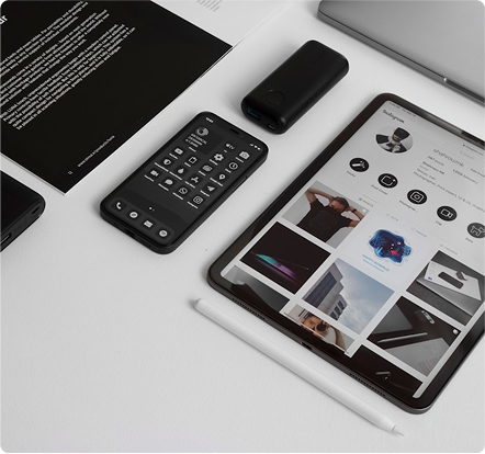 Flat lay of a smartphone with dark mode interface, a black power bank, a tablet displaying an Instagram profile, and a white stylus on a white surface.