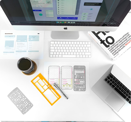 Top view of a white desk with a computer, laptop, design sketches, stencils, pen, and a cup of coffee, suggesting UI/UX or app design work.