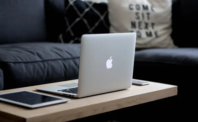 Ein Laptop mit Apple-Logo auf einem Holztisch vor einem Sofa, daneben liegen ein Tablet und ein Smartphone.