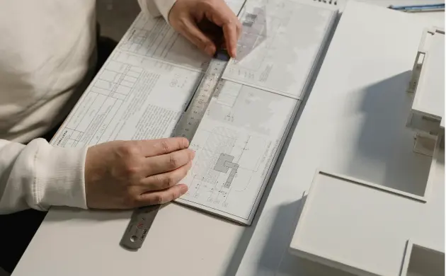 Person misst mit einem Lineal auf technischen Bauzeichnungen auf einem Schreibtisch.