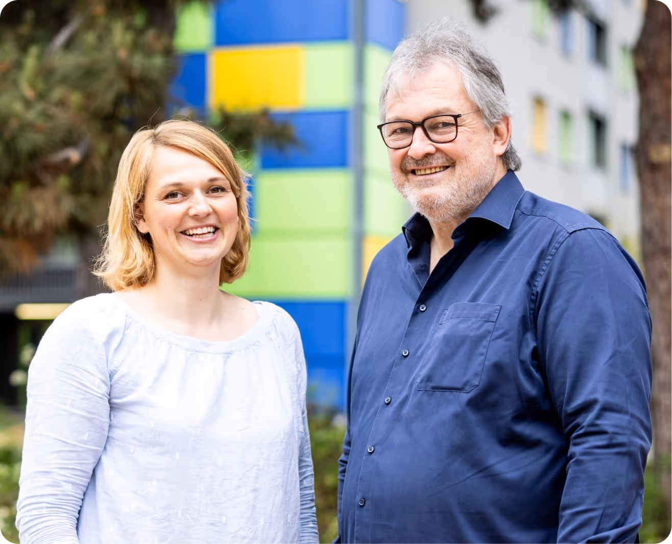 Lächelnde Frau mit blonden Haaren in weißer Bluse und Mann mit grauem Haar, Bart und Brille in blauem Hemd vor einem farbenfrohen Gebäude.