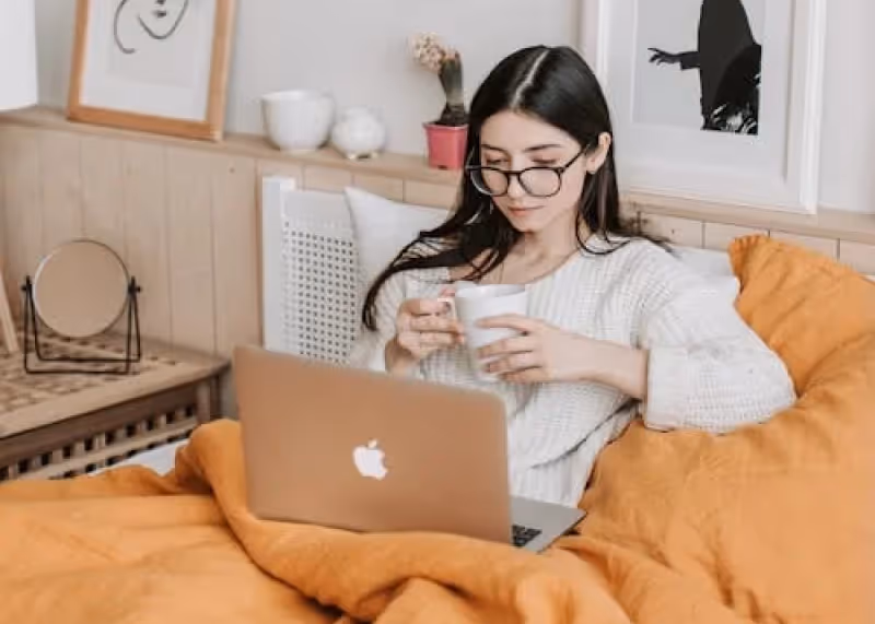 Frau mit Brille sitzt im Bett, trinkt aus einer Tasse und benutzt einen Laptop.
