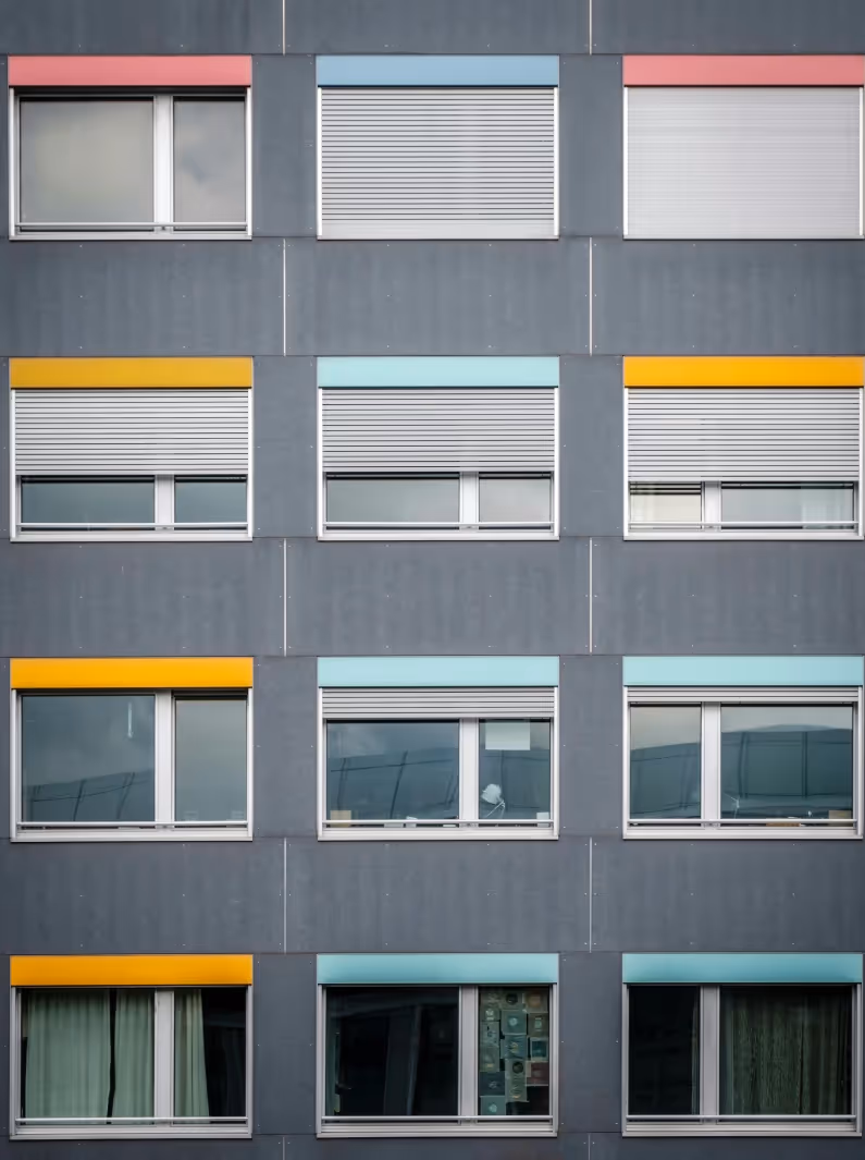 Fassade eines Gebäudes mit zwölf Fenstern in vier Reihen, einige mit bunten Rahmen in Rot, Gelb und Blau und unterschiedlichen Jalousiestellungen.
