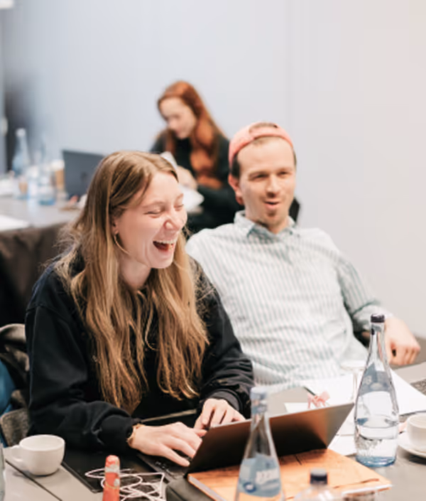 Zwei junge Erwachsene lachen zusammen während sie an einem Laptop an einem Tisch mit Wasserflaschen und Tassen arbeiten.