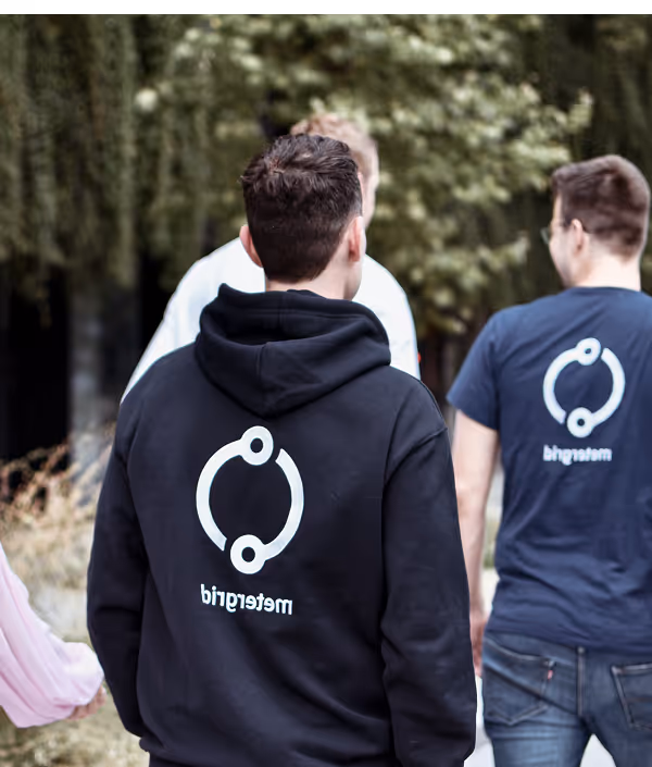 Drei Personen gehen draußen, zwei von ihnen tragen Kleidung mit dem Logo von 'metagrid' auf dem Rücken.