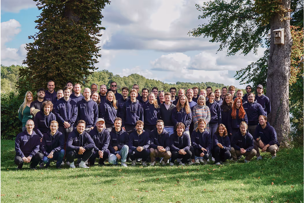 Große Gruppe von etwa 50 Personen, die draußen auf einem grünen Rasen vor Bäumen und bewölktem Himmel posieren.