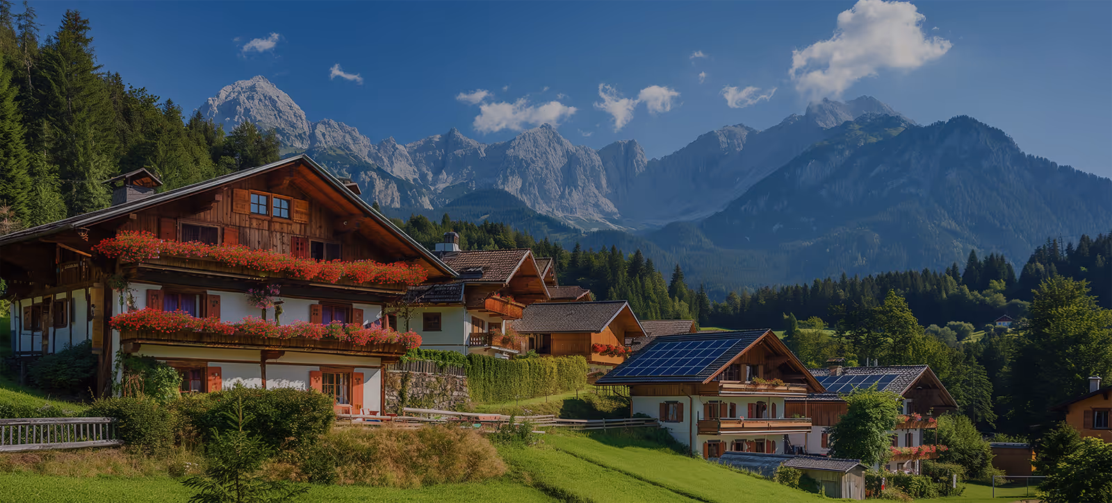 Bayerische Berglandschaft mit traditionellen Häusern, blühenden Balkonen und Solarpaneelen vor bewaldeten Bergen und blauem Himmel.
