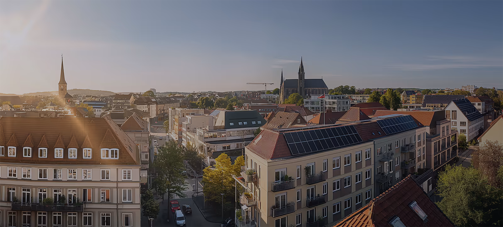 Stadtansicht mit Wohngebäuden, Solarpaneelen auf einem Dach, zwei Kirchtürmen und klarem Himmel bei Sonnenuntergang.