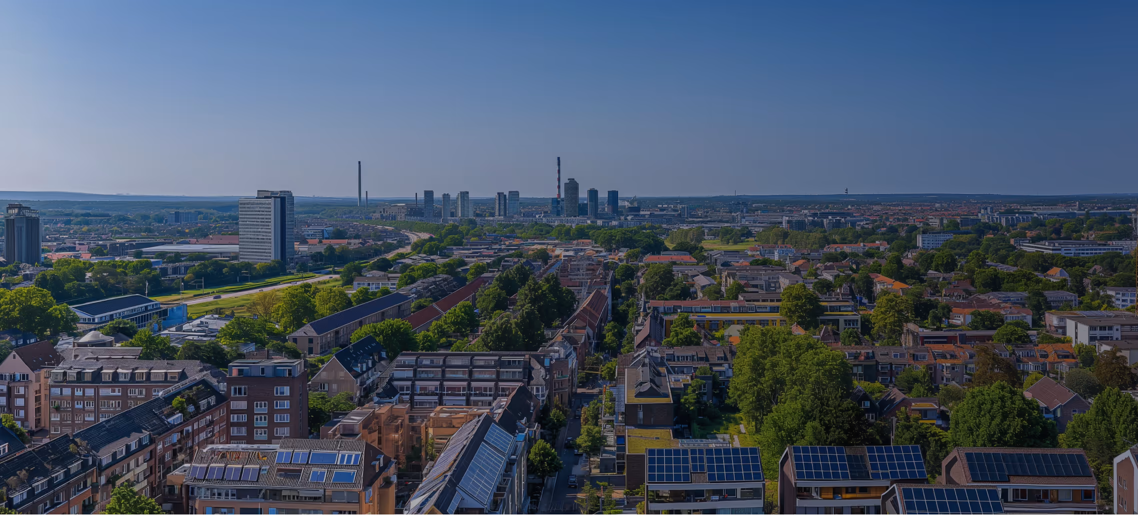 Stadtlandschaft mit Wohnhäusern, vielen Bäumen und Hochhäusern im Hintergrund unter klarem blauem Himmel.