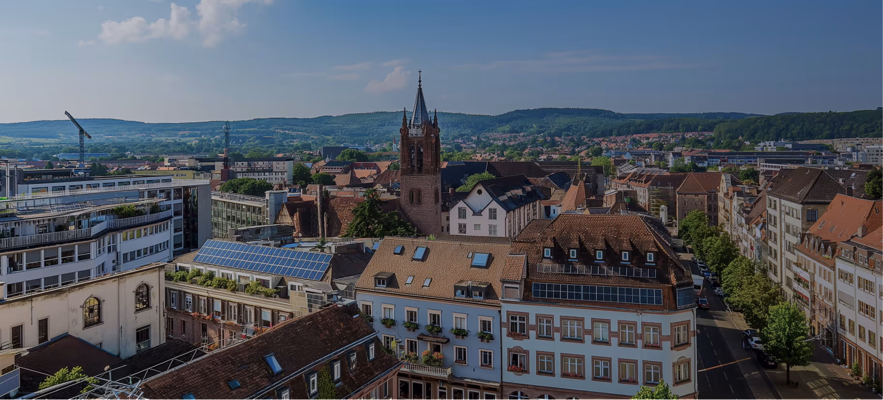 Stadtansicht mit historischen Gebäuden und einem Kirchturm vor bewaldeten Hügeln unter blauem Himmel.