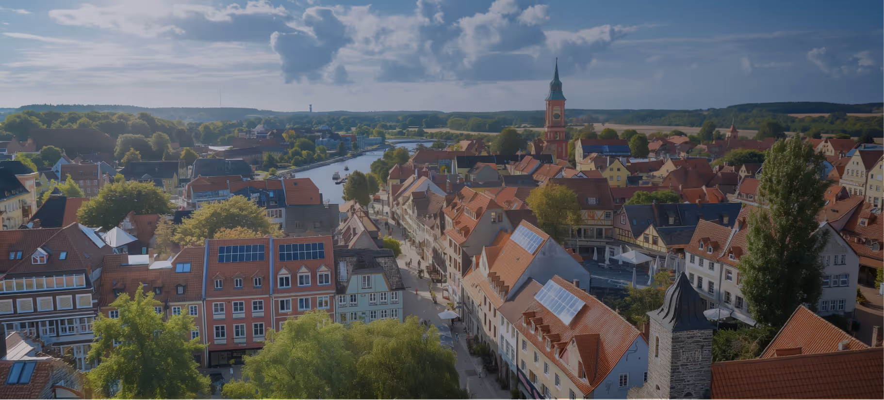 Luftaufnahme einer malerischen deutschen Stadt mit roten Ziegeldächern, einem Fluss und einer Kirchturmspitze bei bewölktem Himmel.