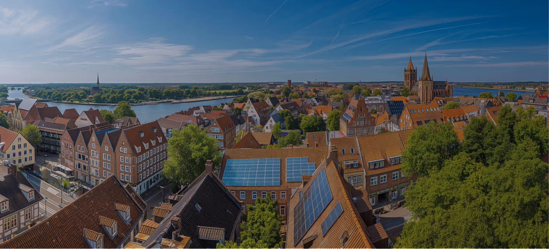 Luftaufnahme der Stadt mit roten Ziegeldächern, Solarpanelen, großen Bäumen, Fluss und historischem Kirchgebäude bei blauem Himmel.