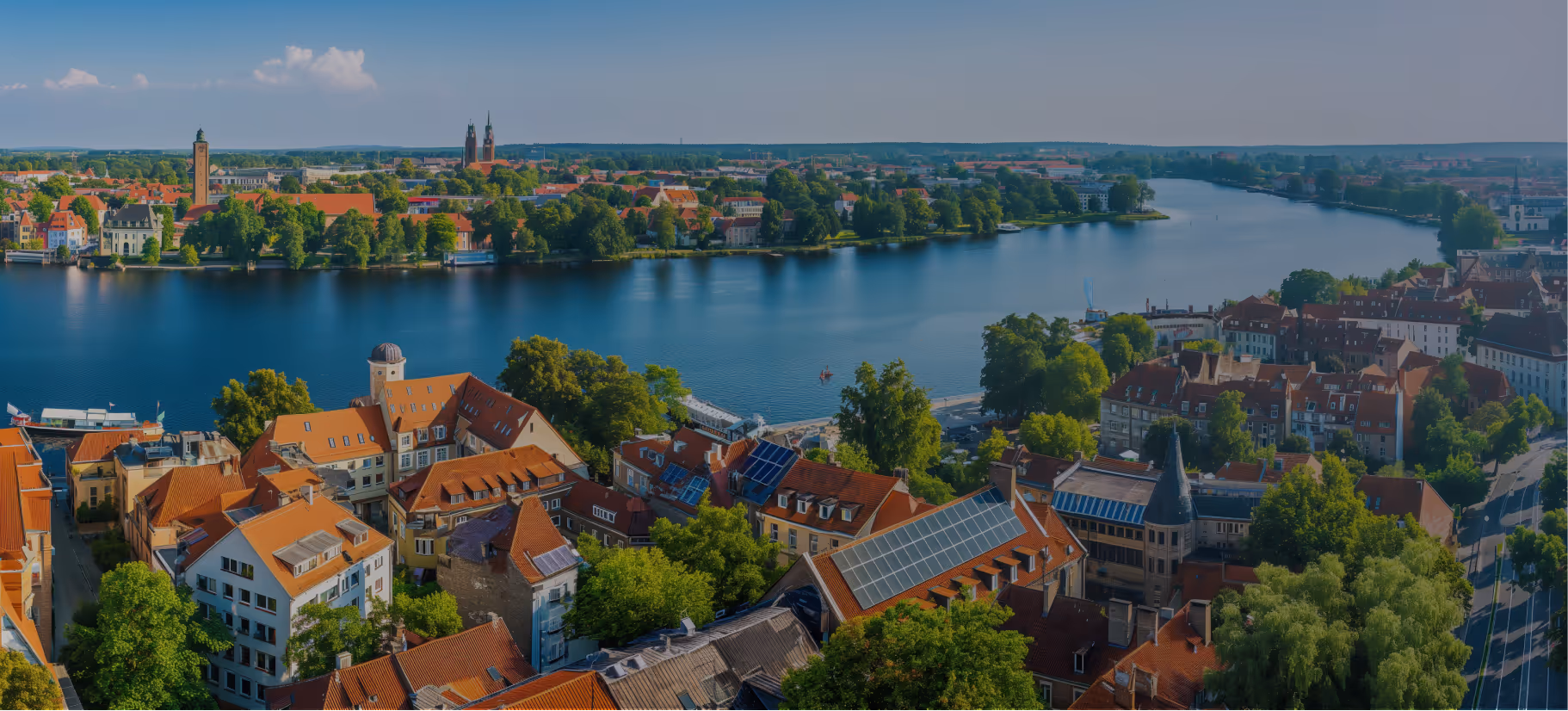Panoramablick auf einen Fluss mit rot gedeckten Häusern und grünen Bäumen an beiden Ufern unter klarem Himmel.