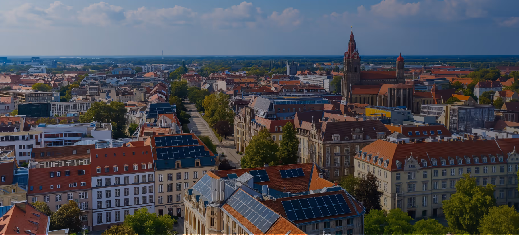 Panoramablick auf eine Stadt mit roten Dächern, mehreren Gebäuden mit Solarpaneelen auf den Dächern und einer großen Kirche im Hintergrund.
