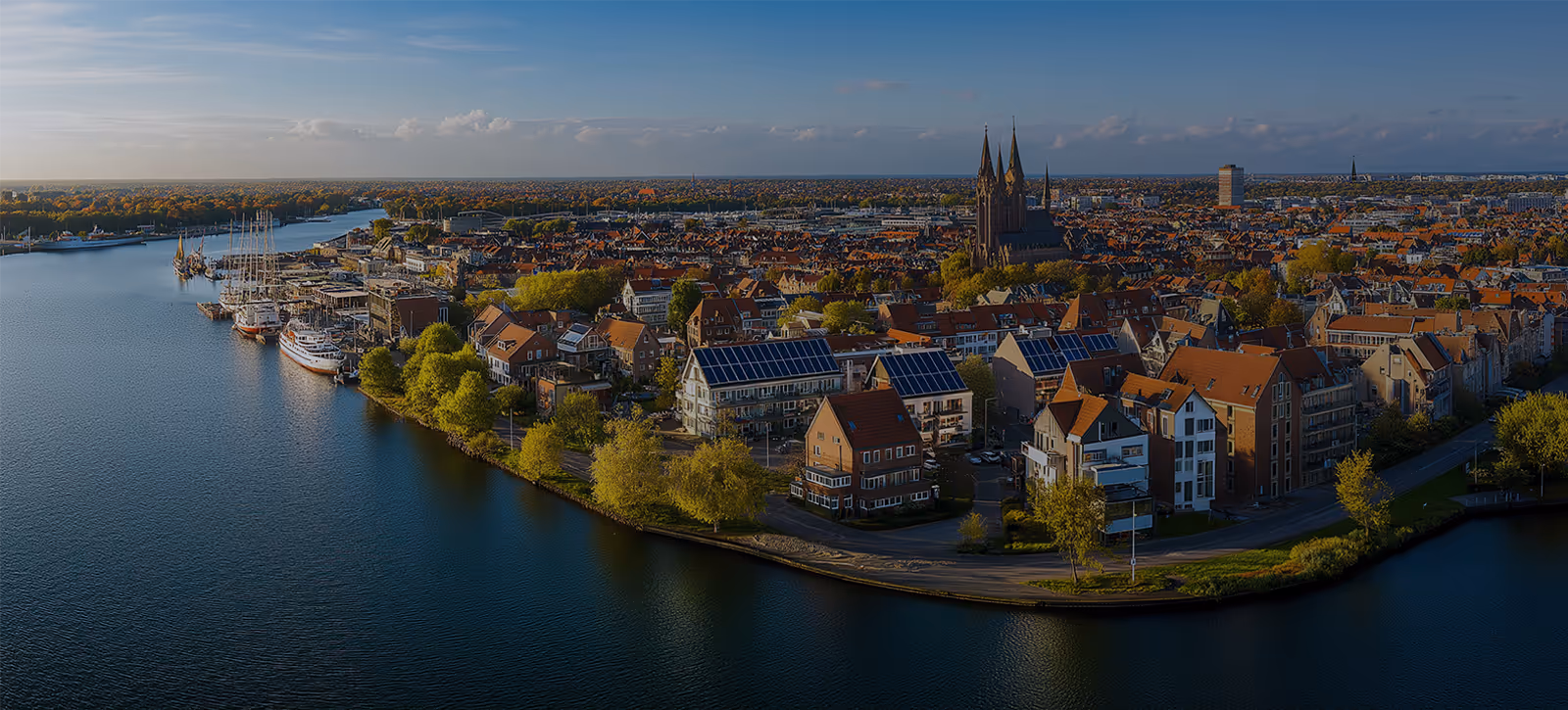 Luftaufnahme einer Stadt mit Wasserweg, Booten, Wohnhäusern mit Solarpanels auf den Dächern und einer großen Kirche im Hintergrund bei klarem Himmel.