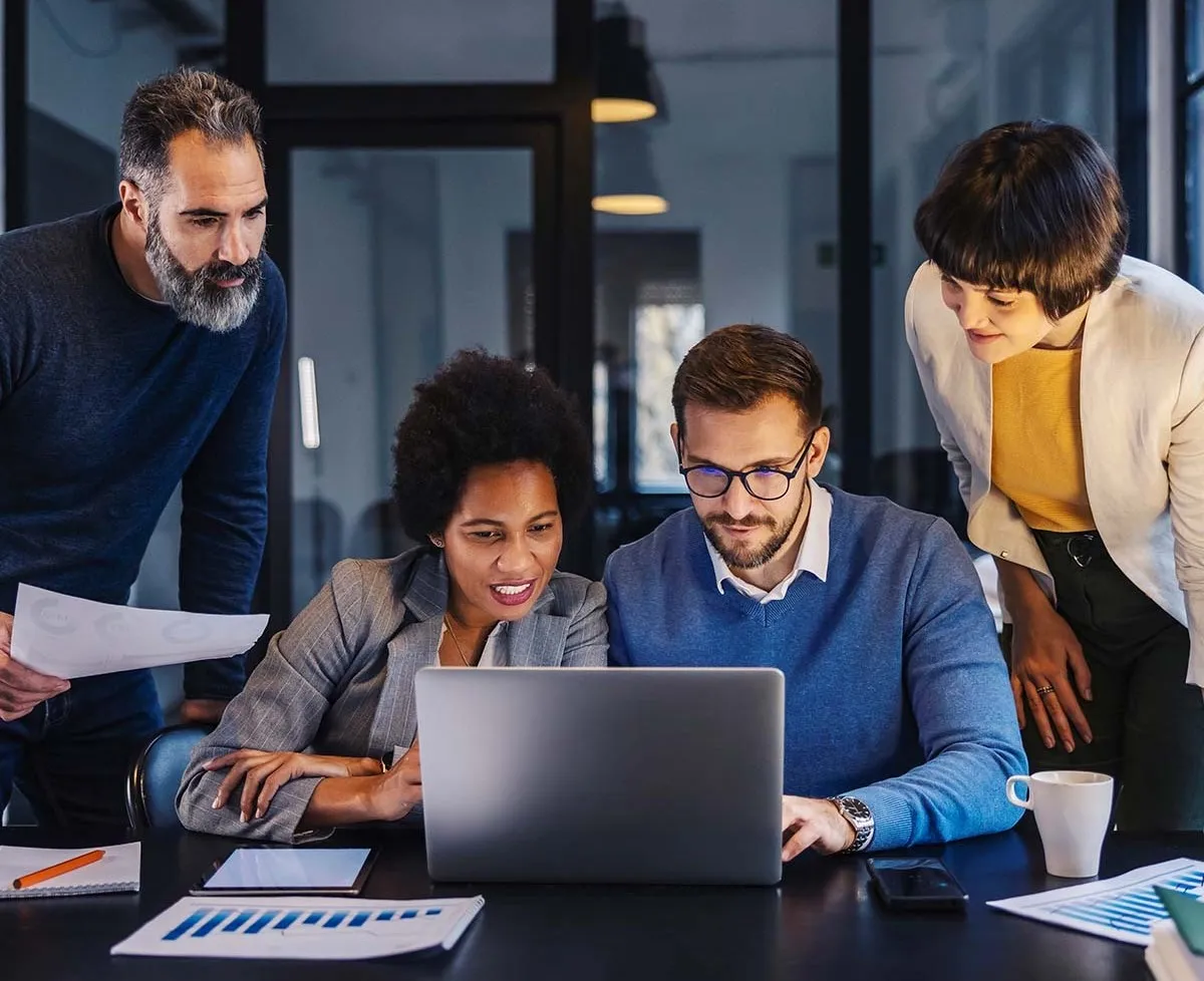 Four colleagues gather around laptop in office, discussing work documents and data charts.