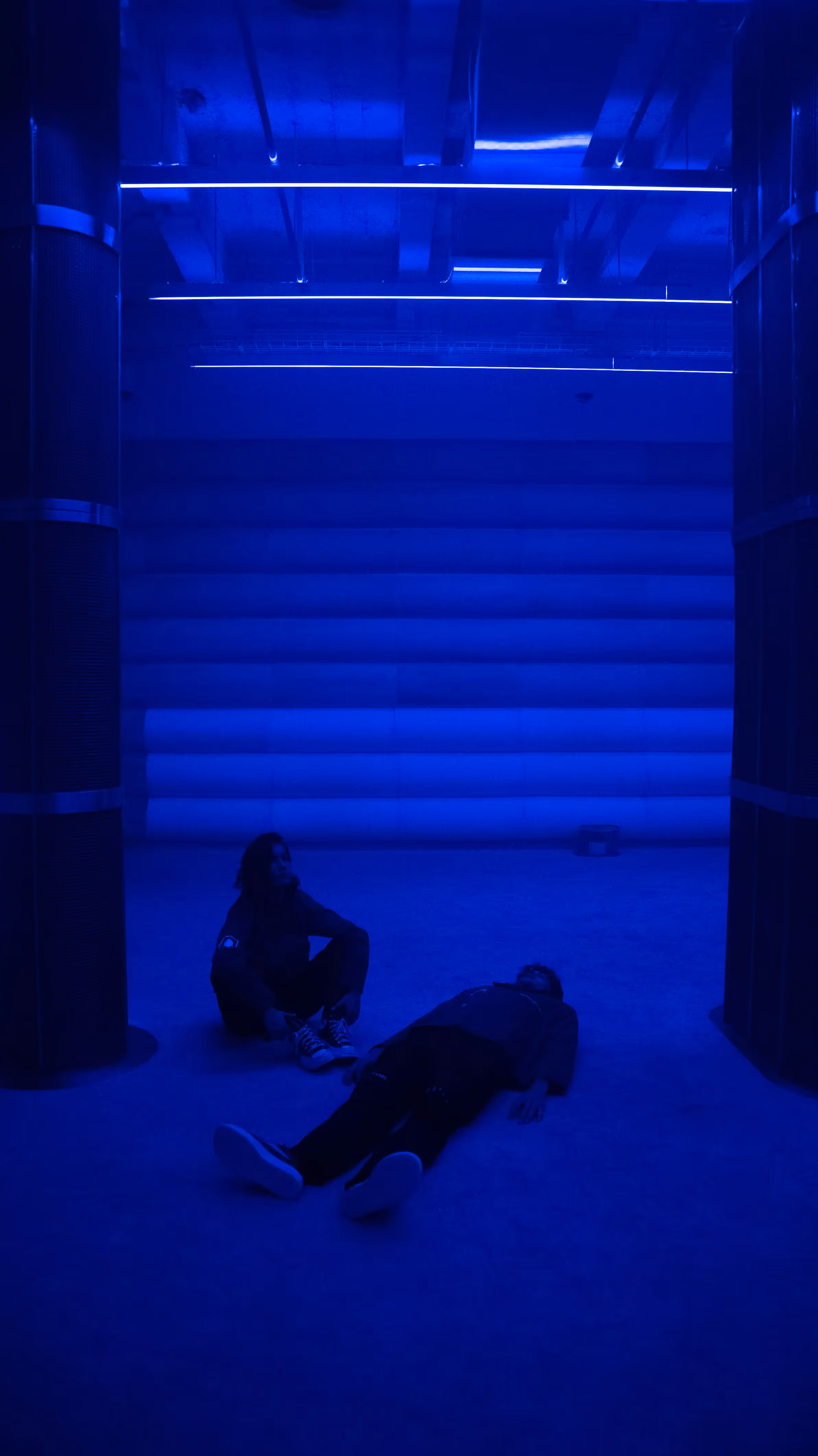 Two visitors rest on the floor of the 4DSound chamber at Mercer Labs, one lying down and one seated, under deep blue light and ceiling LED strips, Lower Manhattan.